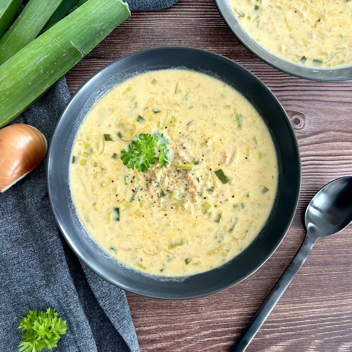 Das Bild zeigt in der Mitte des Bildes in einer schwarzen tiefen SChüssel die Käselauchsuppe. Rechts davon liegt ein Esslöffel. Die Suppe ist mit Petersilie verfeinert.