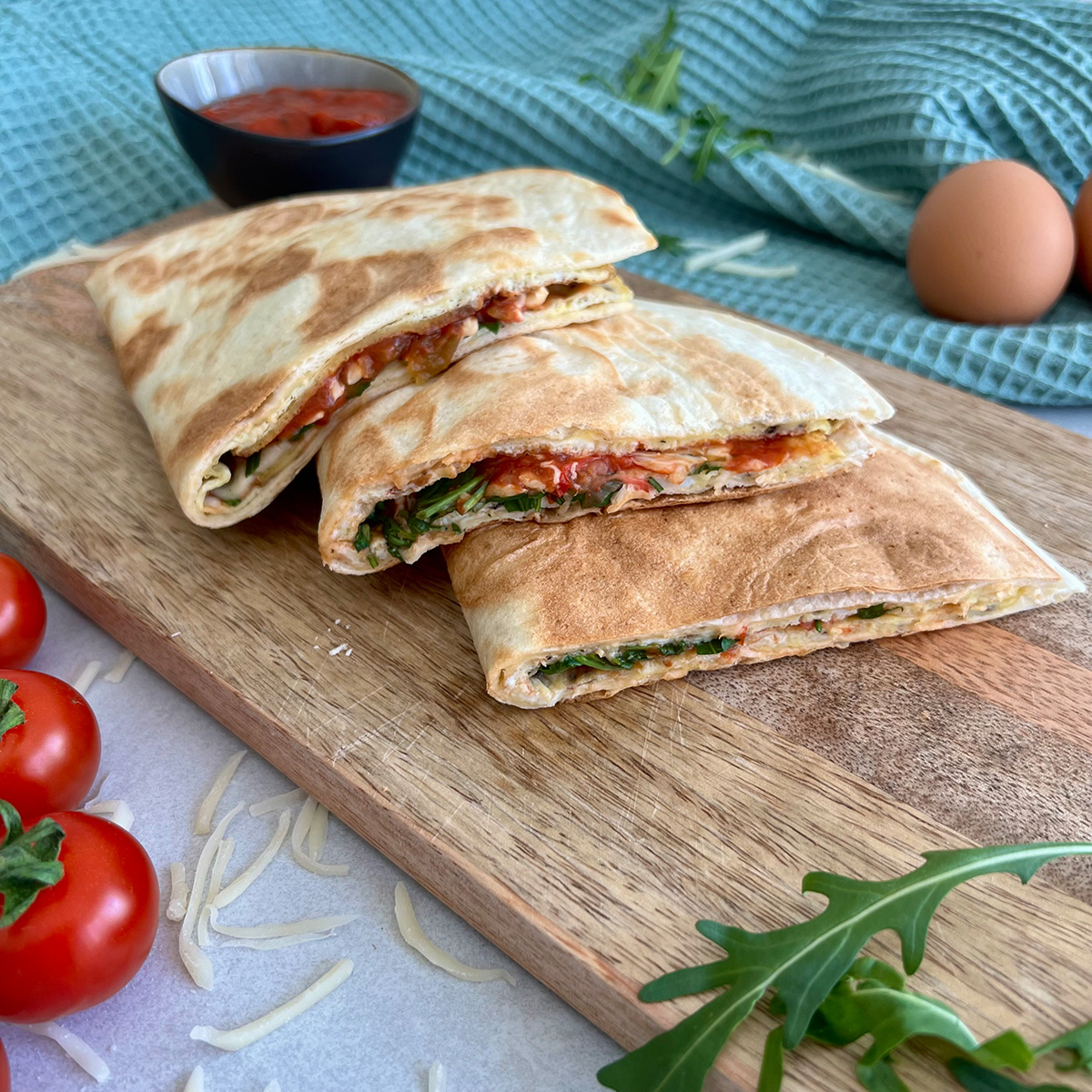 FrühstückswrapsDas Bild zeigt 3 Wraps übereinanderliegend auf einem Brettchen. Ruccola, Tomaten und Eier liegen daneben und ein Dip steht bereit.