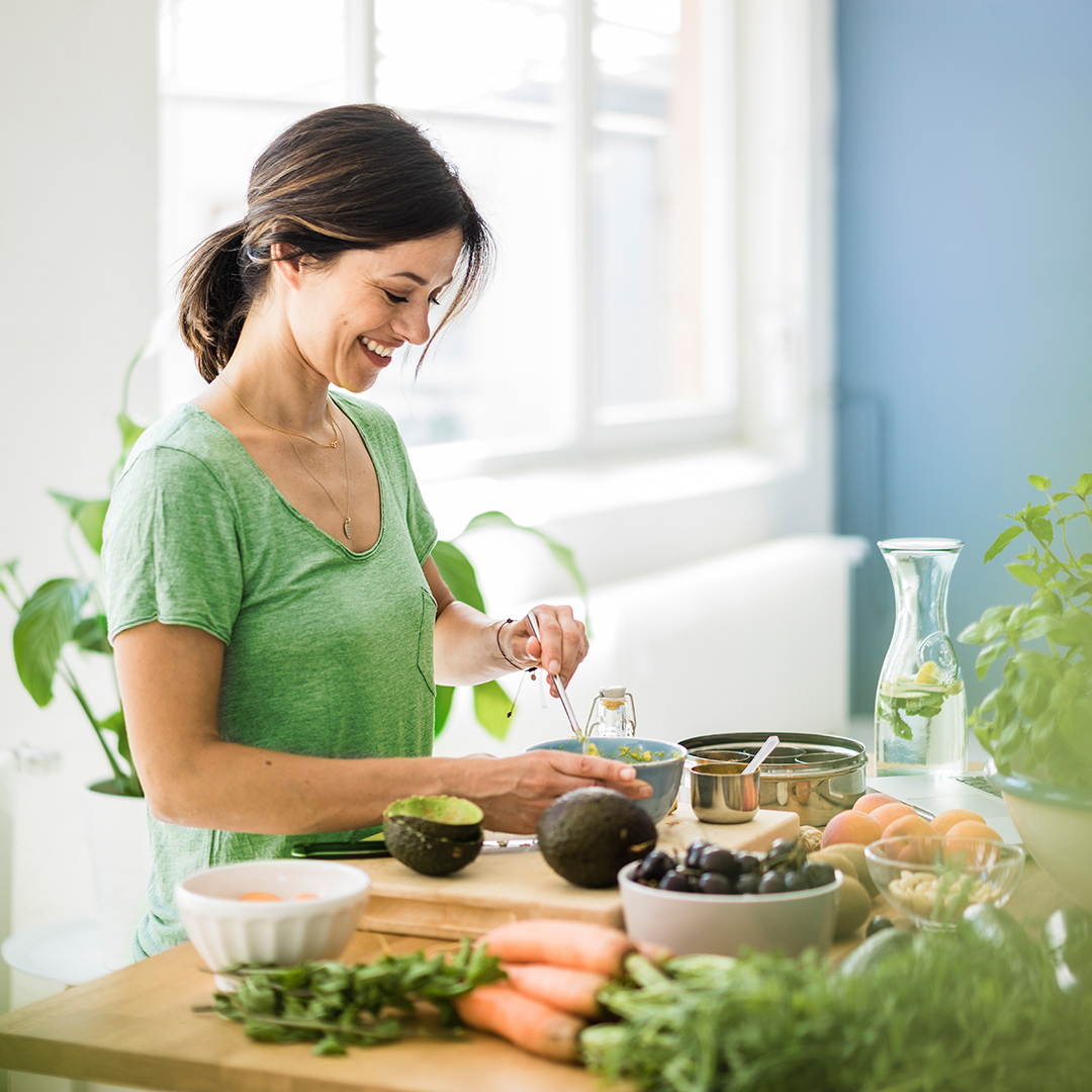 Eine lächelnde Frau mit grünem T-Shirt, bereitet in heller Küche  umgeben von frischem Gemüse und Kräutern, eine Mahlzeit zu.