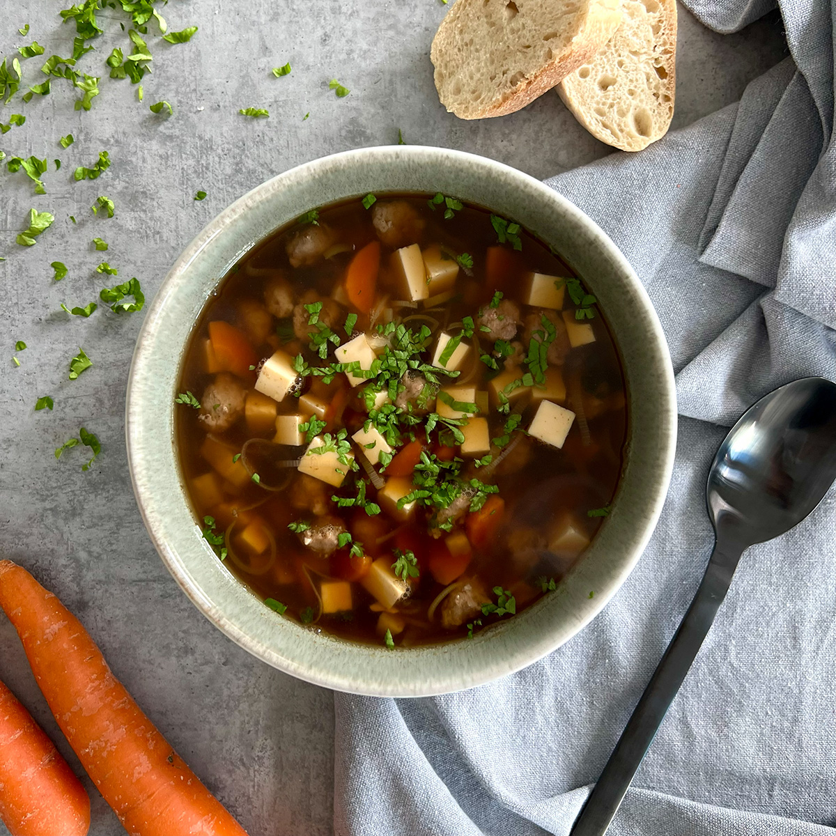 Eine Schüssel mit besagtem Gericht, deutlich zu sehen ist Eierstich und die Brühe. Nebenbei Brot, Karotten, Kräuter und ein Löffel