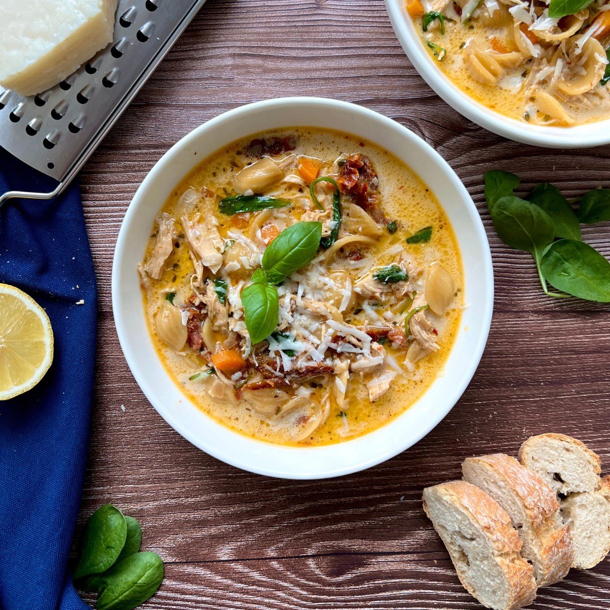Das Bild zeigt eine toskanische Hähnchen Suppe in einer weißen tiefen Schüssel getoppt mit Basilikum. Neben der Schüssel sind Baguette Scheiben eine Käse Reibe und Parmesan zu sehen.