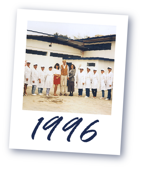 Eine Gruppe von Menschen, darunter mehrere in weißen Kitteln, vor einem Gebäude. Darunter die Jahreszahl '1996' darunter