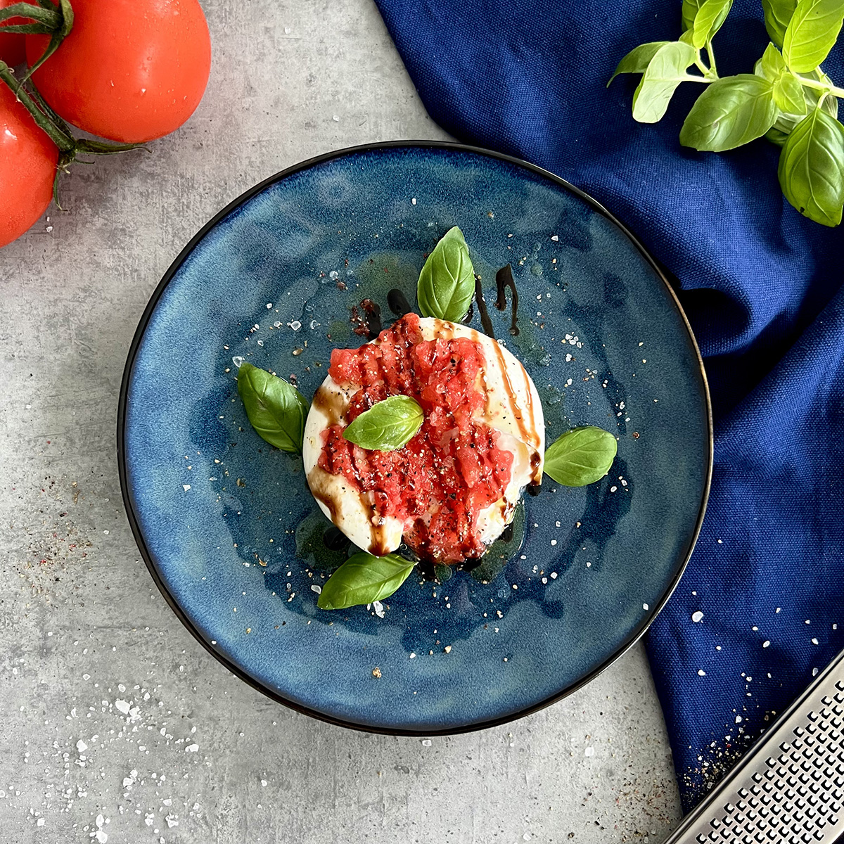 Eine Burrata Kugel getoppt mit geriebener gefrorener Tomate, Balsamico Creme, Gewürzen und Basilikum auf einem runden Teller.