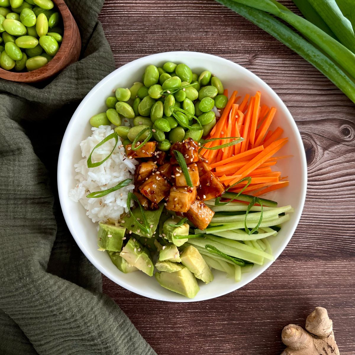 Das Bild zeigt eine Bowl in einer weißen tiefen Schüssel. Daneben stehen Edamame, Frühlingszwiebeln und Ingwer.