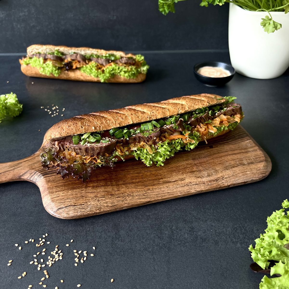 Ein Sandwich belegt mit Salat, Kimchi, Steakscheiben und Frühlinsgzwiebel auf einem Holzbrett. Im Hintergrund ein weiteres Sandwich.