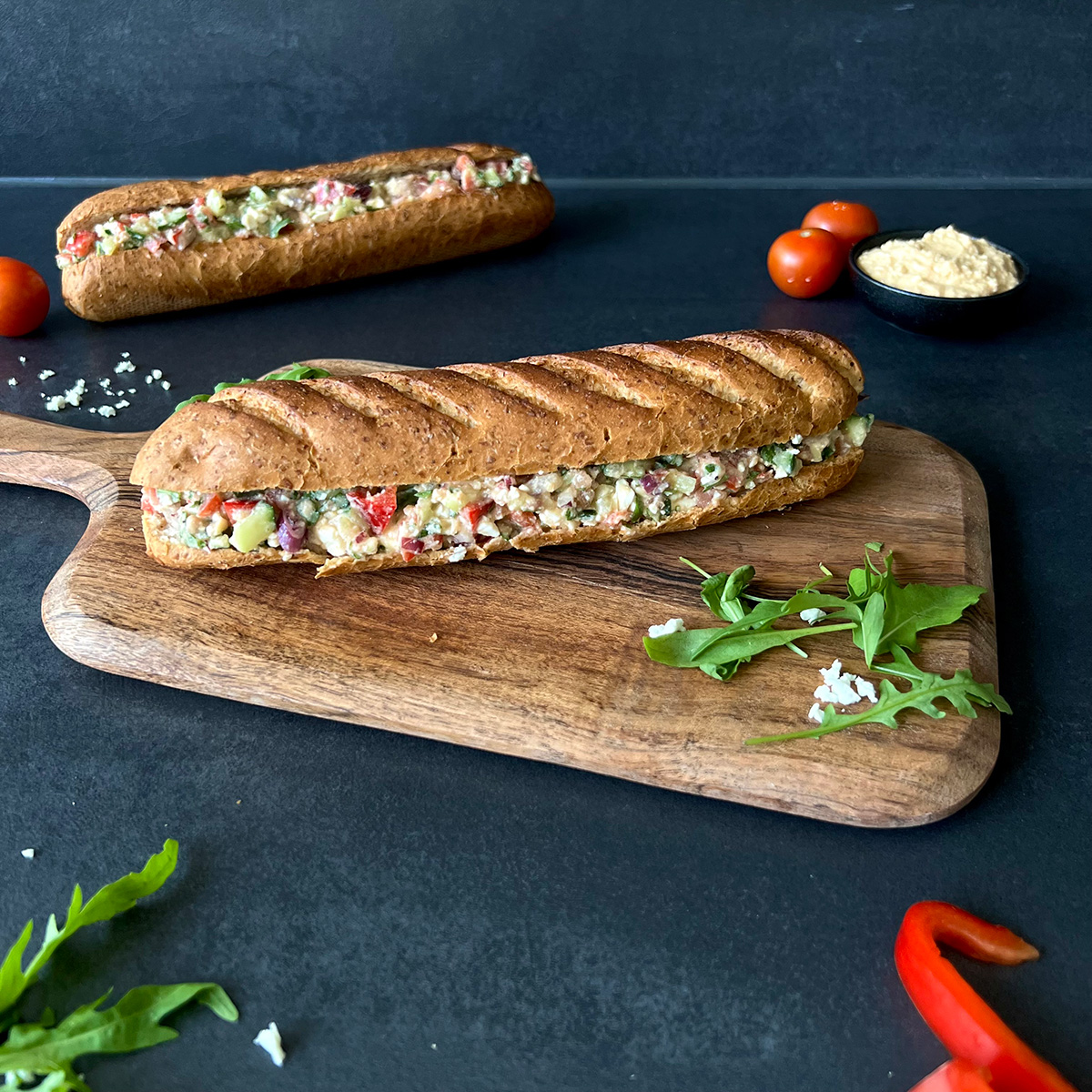 Das Bild zeigt ein Sandwich auf einem Brettchen. Ein weiteres liegt im Hintergrund. Paprika, Salat und Hummus ist daneben serviert.