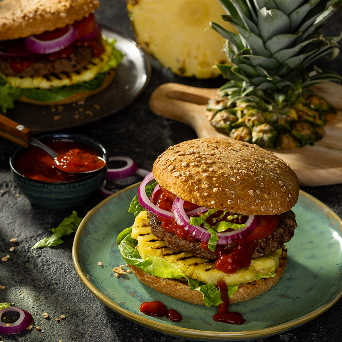 Ein Burger mit gegrillter Ananas, Patty, Salat, Sauce und Zwiebelringen auf einem Teller. Im Hintergrund eine aufgeschnittene Ananas.