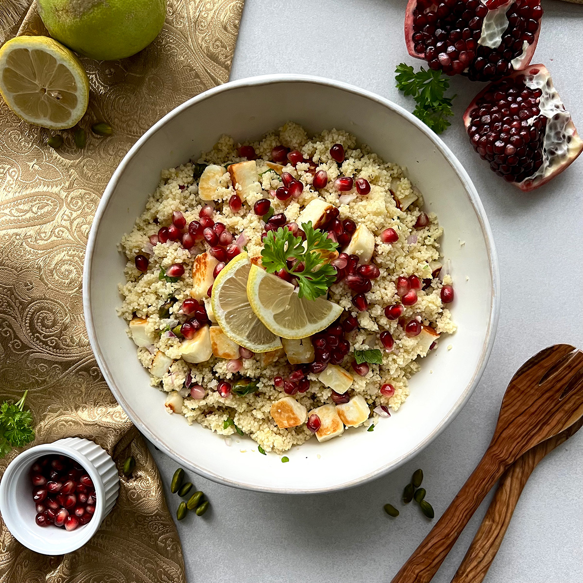 Das Bild zeit eine weiße Schale gefüllt mit Couscous getoppt mit Halloumi und Granatapfelkernen. Ein Granatapfel und Salatbeteck liegt daneben.