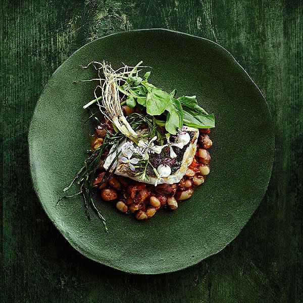 Ein Gericht auf einem dunkelgrünen Teller. Im Mittelpunkt liegt ein Stück gebratenen Fisch, der auf einem Bett aus roten und weißen Bohnen ruht. Der Fisch ist mit verschiedenen Kräutern und essbaren Blüten dekoriert. 
