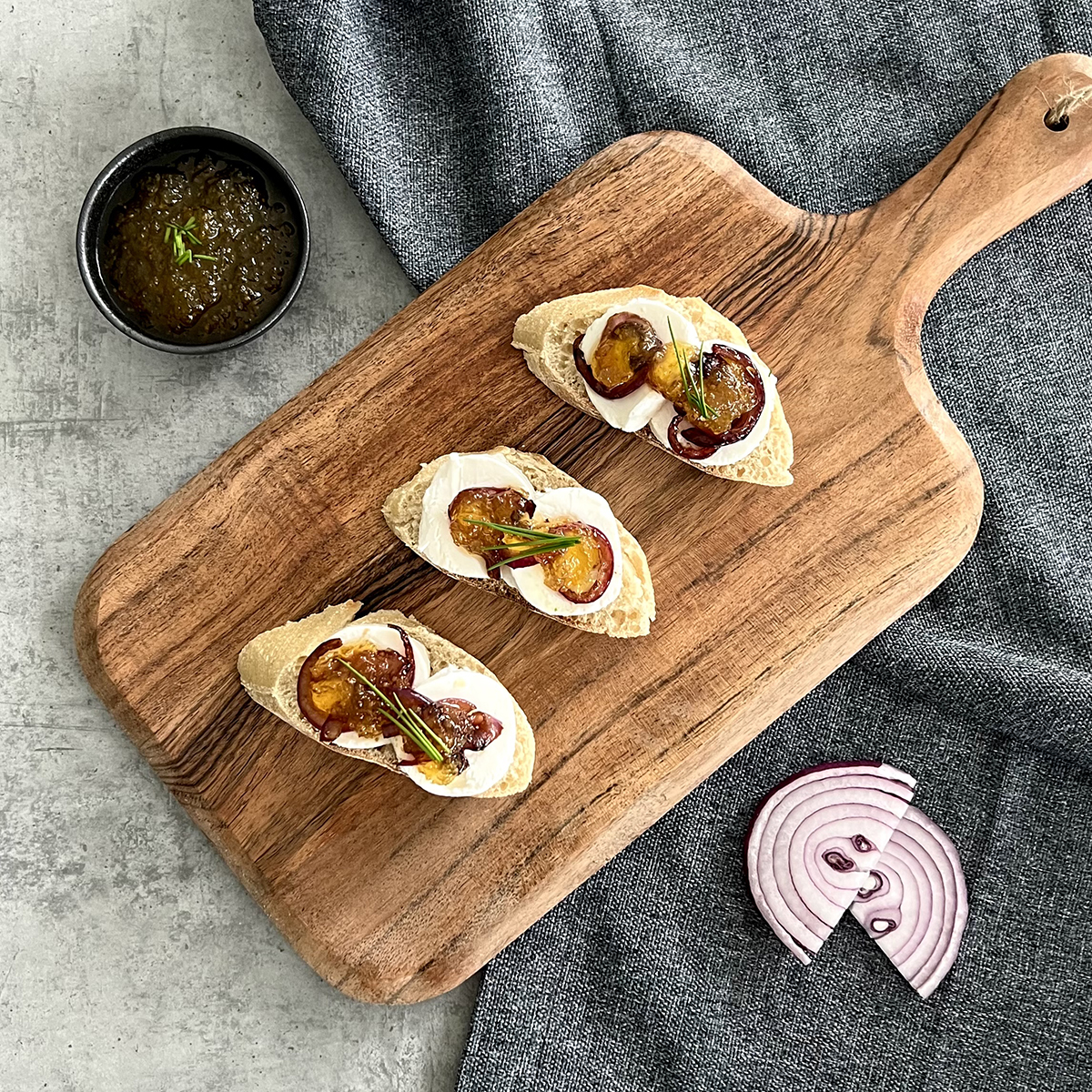 Das Bild zeigt 3 Baguettescheiben belegt mit Ziegenkäse, Zwiebeln und Feigen-Senf-Sauce auf einem Holzbrett.