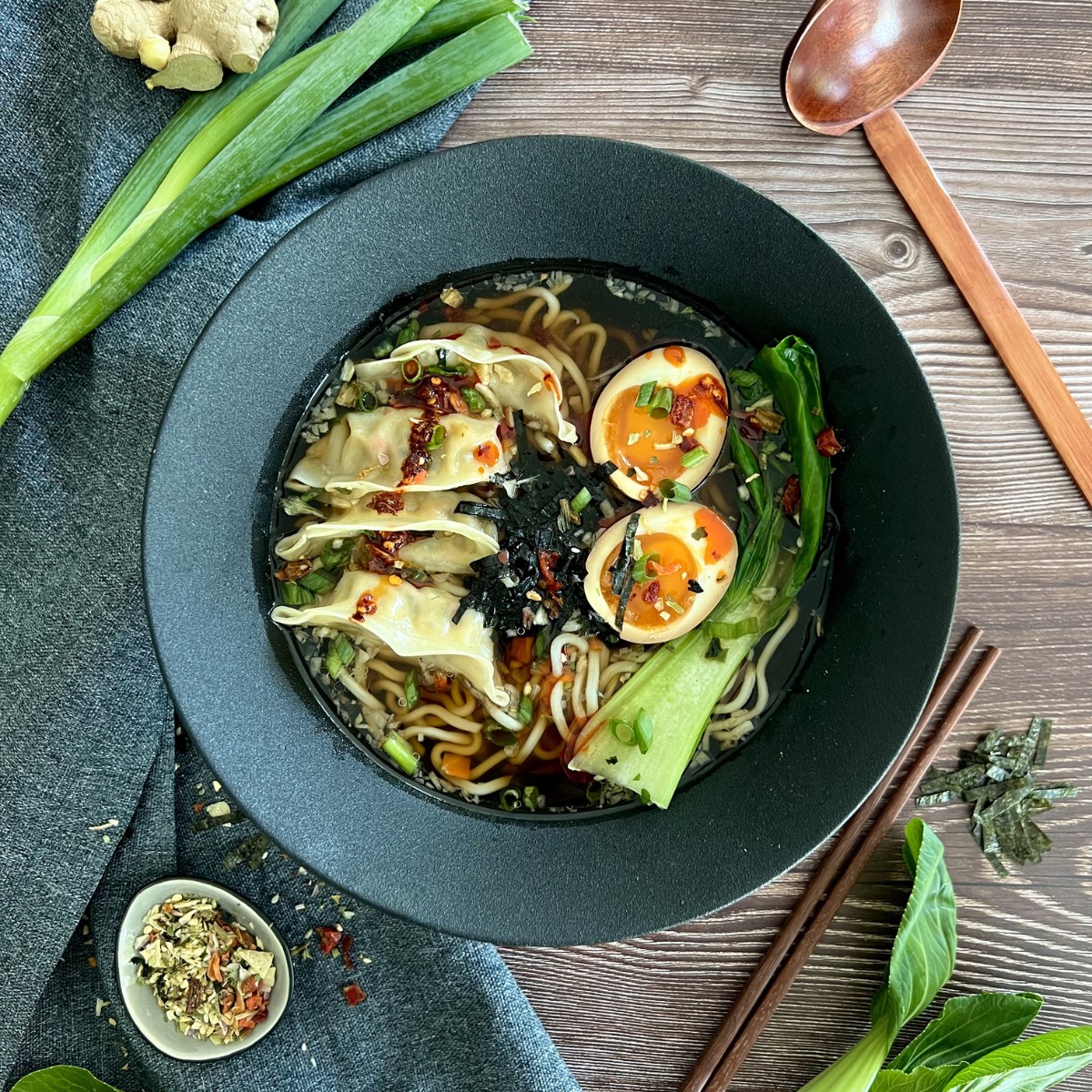 Die Ramen ist in einer schwarzen tiefen Schüssel serviert. Daneben sind die Gewürze in einem kleinen Schälchen, Frühlingszwiebeln, Ingwer und Essstäbchen.