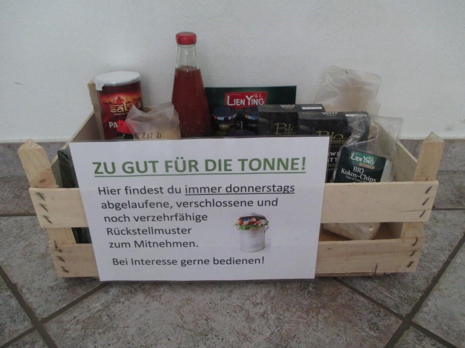 Holzkiste mit abgelaufenen, verschlossenen Lebensmitteln und Schild 'Zu gut für die Tonne! .