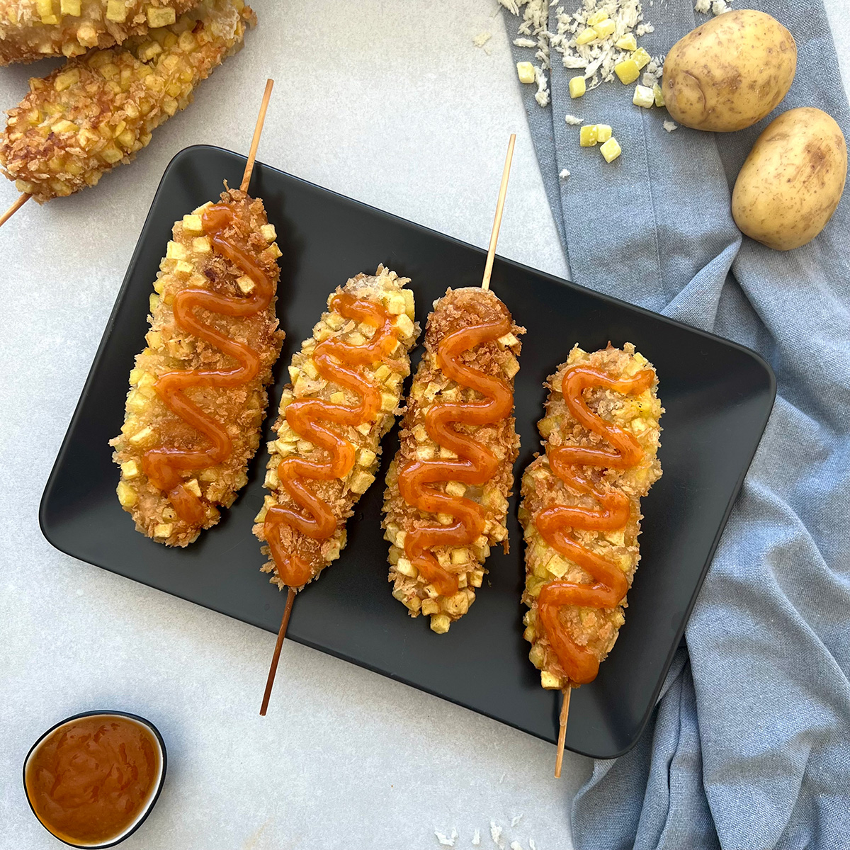 Das Bild zeigt  4 Corndogs auf einem dunklen Teller. Zwei weitere werden angeschnitten und es stehen Kartoffeln und Dip dabei.