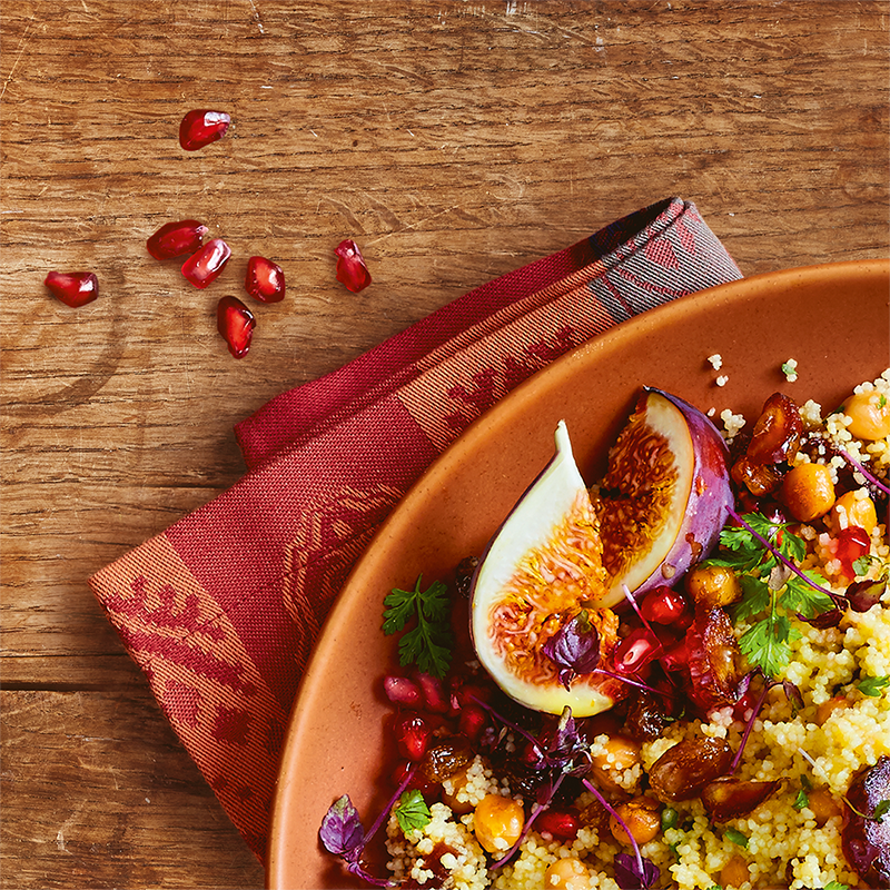 Ein gedeckter Teller symbolisch für die Ethno-Küche mit Couscous, Feigen, Datteln, Kichererbsen und Granatapfelkernen auf einem rustikalen Holztisch.