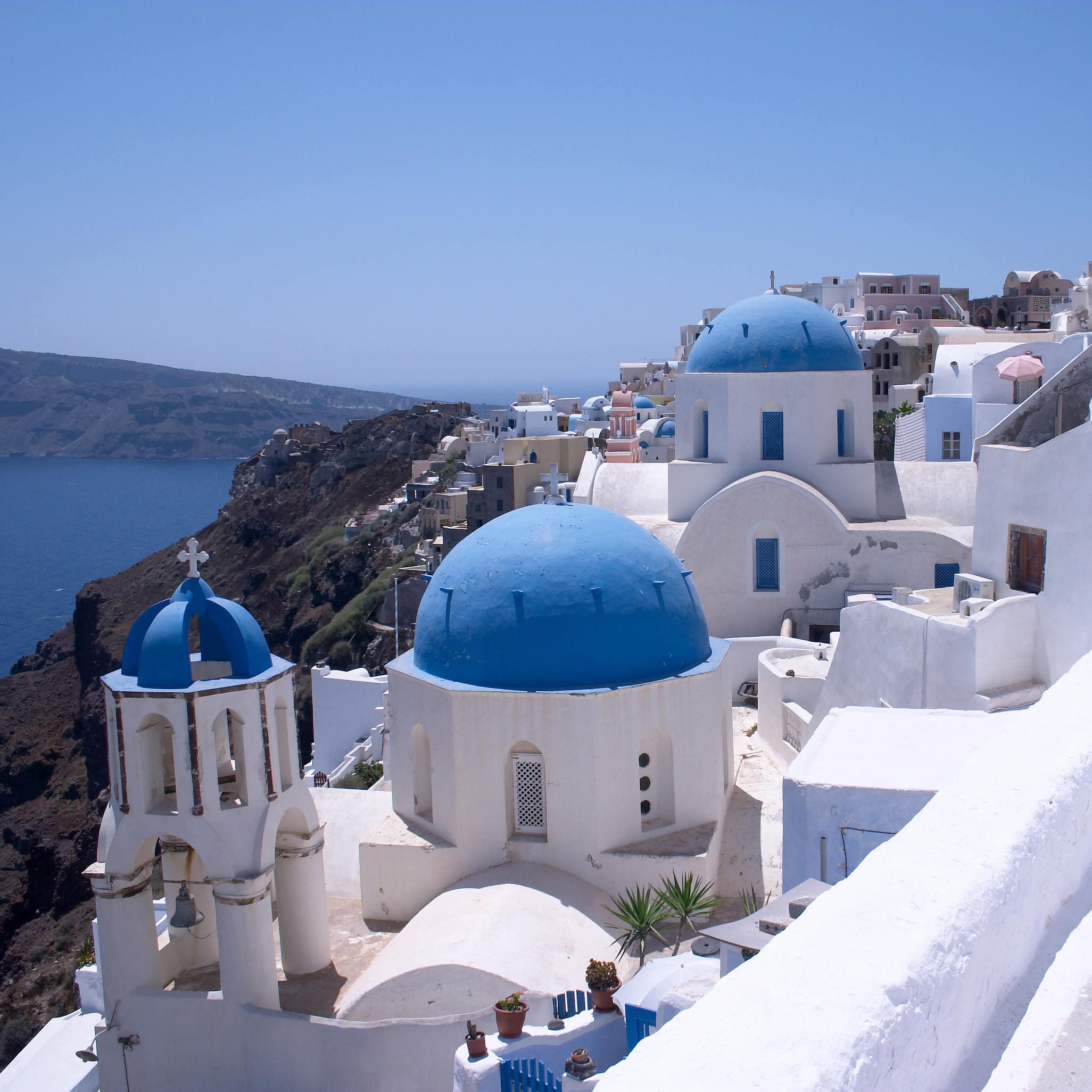Weiße Gebäude mit blauen Kuppeln und Glockenturm auf einer Klippe mit Blick auf das Meer in Griechenland.