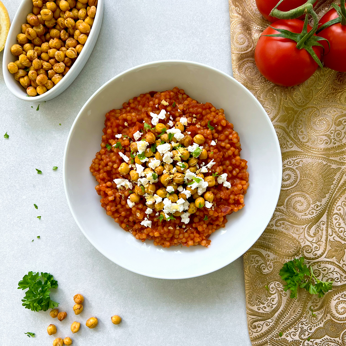 Das Bild zeigt eine Schale mit Tomaten Perlcouscous, der mit knusprigen Kichererbsen und zerbröseltem Feta garniert ist.