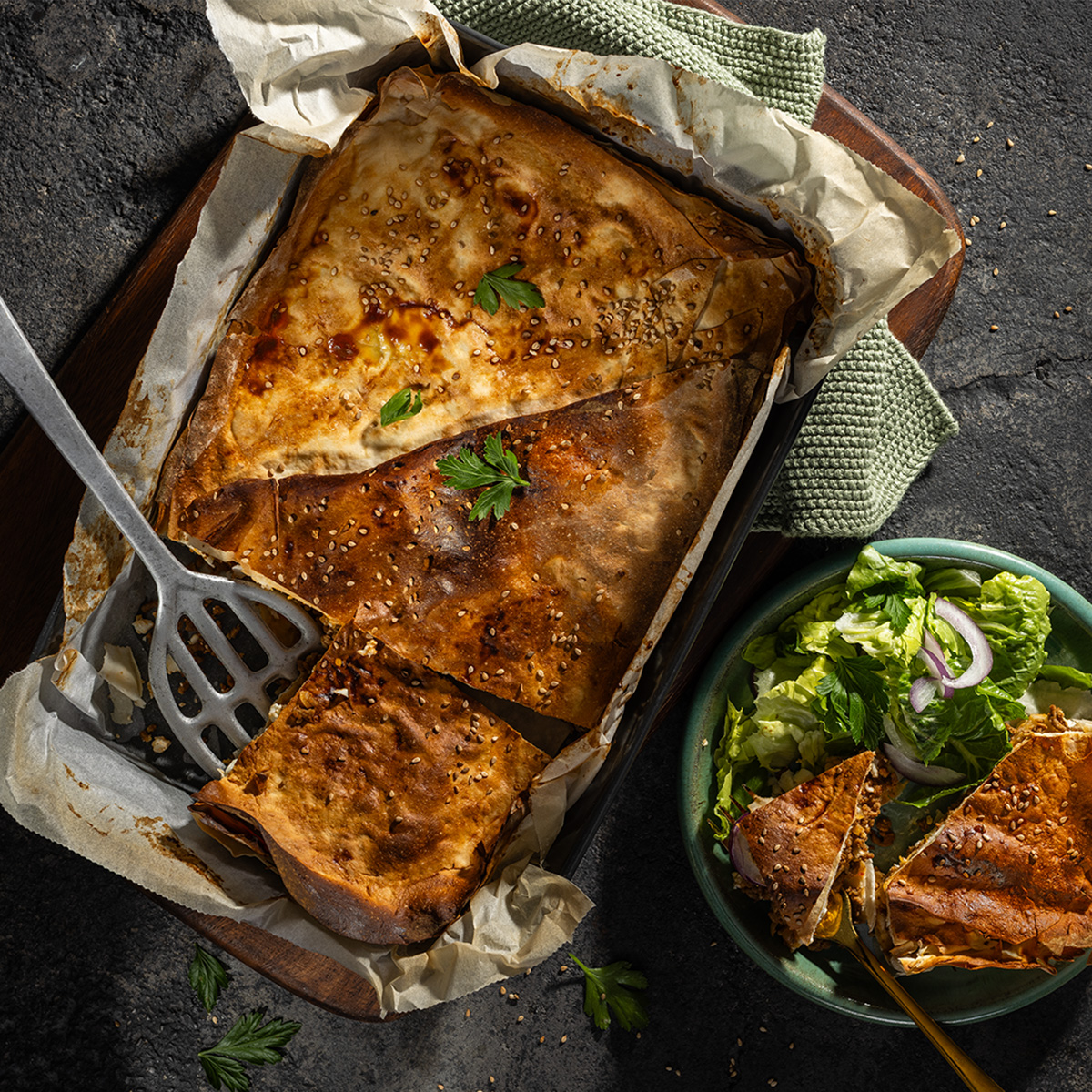 das Bild zeigt Börek auf einem kleinen Blech. Ein Teller mit einem Stück Börek und Salat steht daneben.