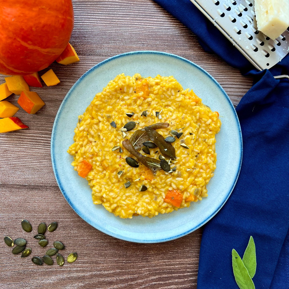 Ein Kürbis Risotto ist flach verteilt auf einem blauen Teller getoppt mit gerösteten Salbeiblättern. Daneben liegt ein Kürbis und Parmesan.