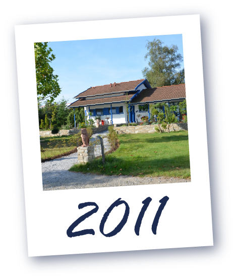 Ein Haus mit rotem Dach und blauen Fensterläden, umgeben von Garten und Bäumen, darunter die Jahreszahl  '2011'.