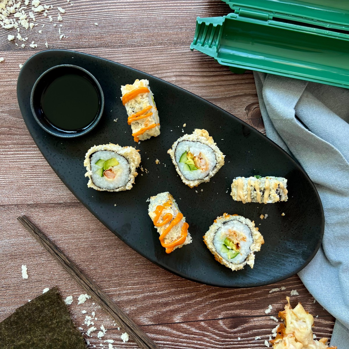 Auf einem länglichen Teller sind sechs Sushistücke verteilt. Die Füllung ist erkennbar und manche sind mit Sriracha Mayo getoppt. Dazu wird Sojasauce serviert.