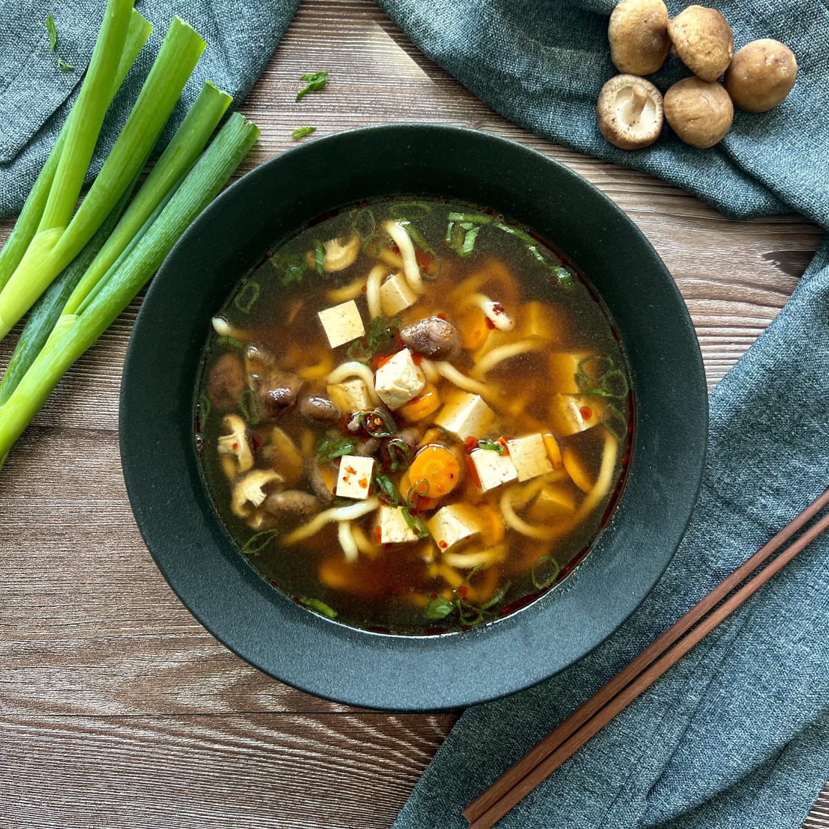 Das Bild zeigt die Miso Udon Suppe in einer schwarzen tiefen Schüssel, sie ist mit Frühlingszwiebeln verfeinert. Außerhalb befinden sich zur Deko Shitake Pilze und Frühlingzwiebeln. Rechts von der Schüssel liegen die Essstäbchen.