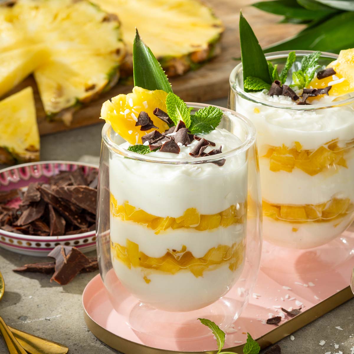Das Bild zeigt zwei Gläser die abwechselnd mit Ananas und Creme geschichtete sind. Serviert werden sie mit Minze und kleinen Schokostückchen.