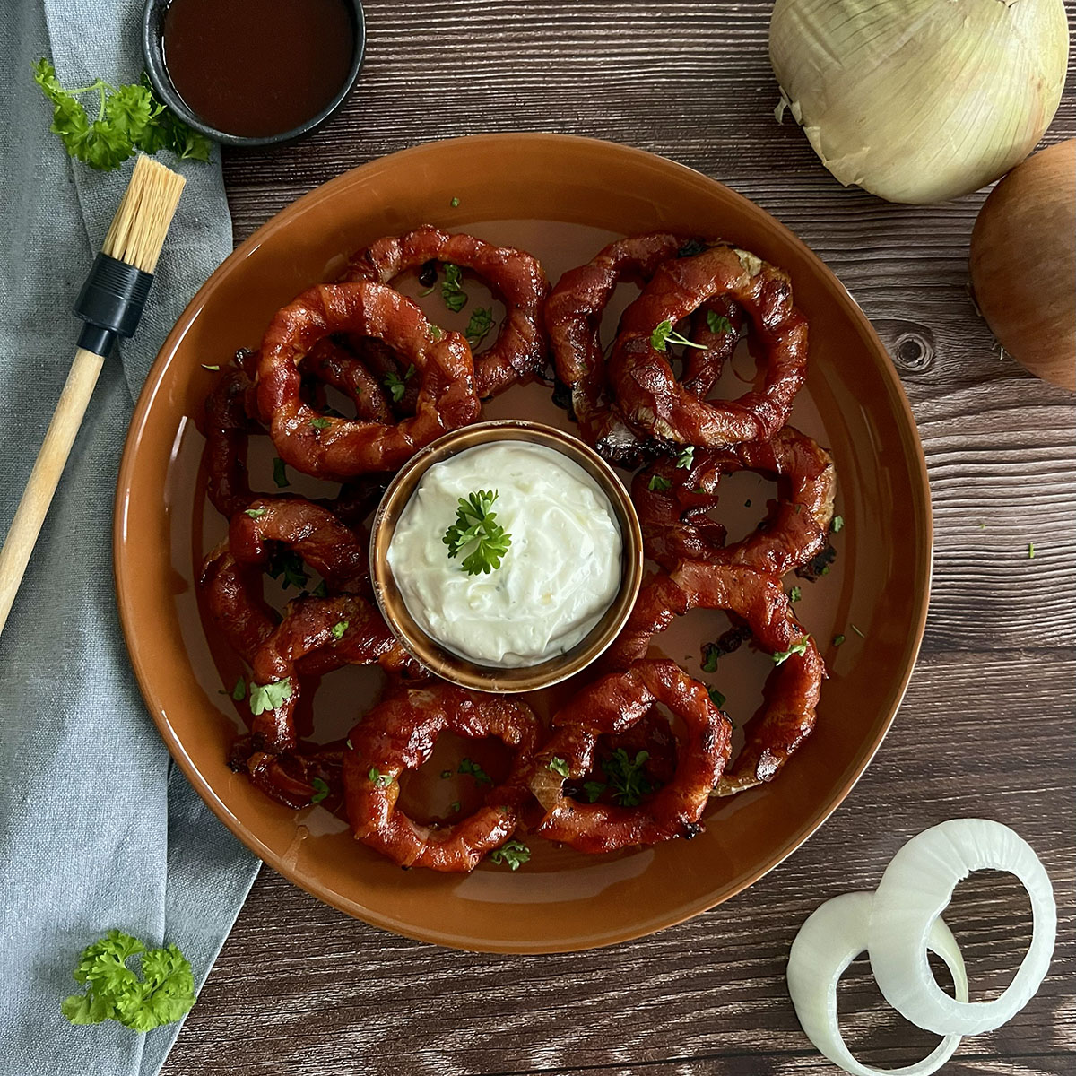 Viele Bacon Zwiebelringe auf einem Teller, in der Mitte ein Dip und am Rand Zwiebeln