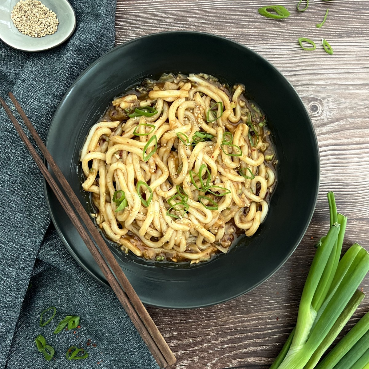 Die Udon sind in einer schwarzen Schüssel darauf liegen zwei Stäbchen. Das Gericht ist mit Frühlingszwiebeln verfeinert.