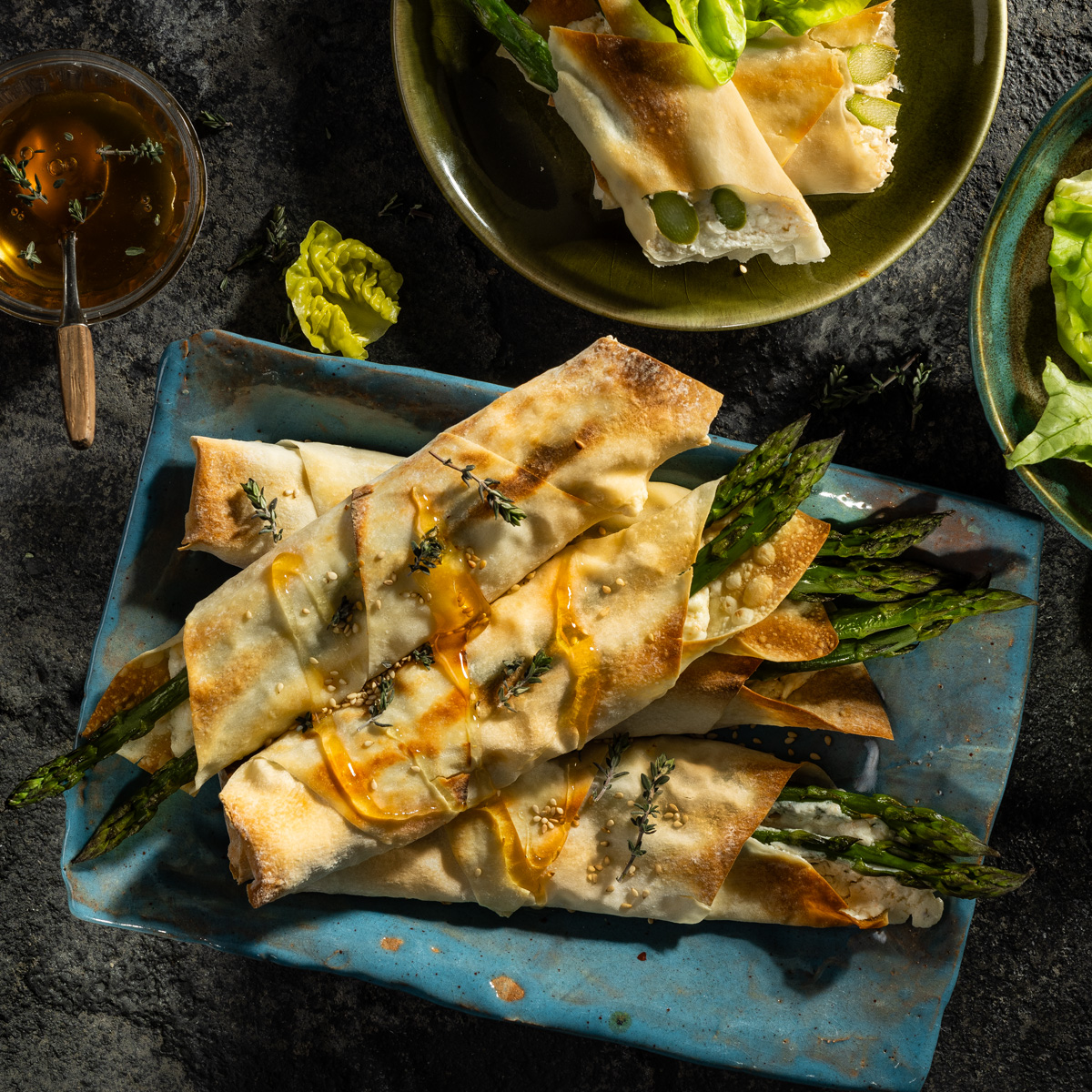 Spargel eingewickelt in gebackenen Yufka Rollen liegen auf einem blauen länglichen Teller. Getoppt mit Honig.
