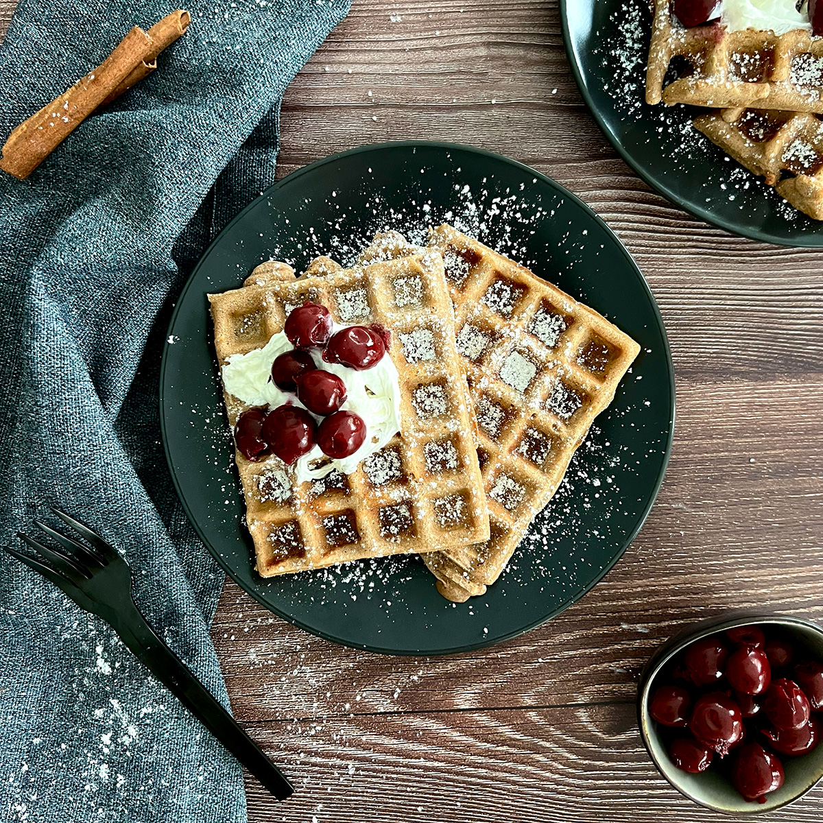 2 Waffeln auf einem Teller garniert mit Puderzucker, daneben weitere Waffeln, Zimt und heiße Kirschen