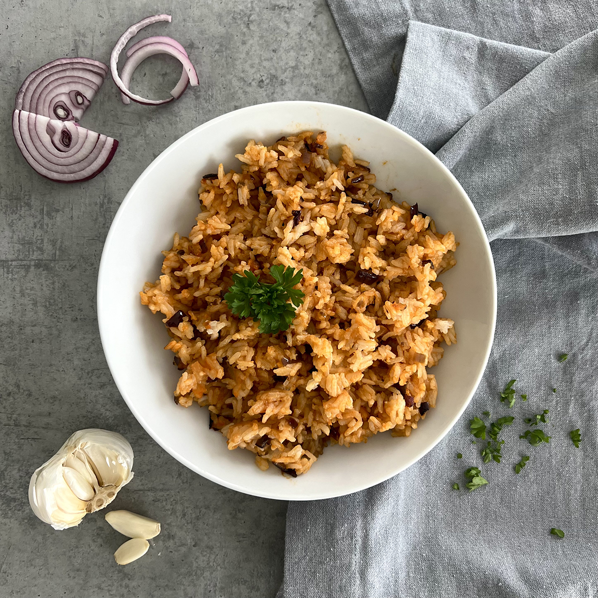 Das Bild zeigt eine weiße Schale mit oranglichen Reis mit Zwiebeln drin. Daneben liegt Knoblauch und Zwiebel. 