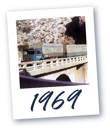 Lastwagen mit zwei Containern auf einer Brücke vor felsigem Hintergrund, darunter die Jahreszahl 1969.