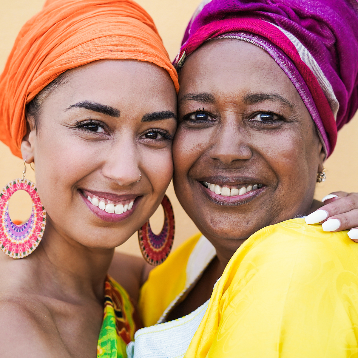 Zwei lächelnde Frauen mit bunten Kopftüchern, die sich umarmen. Eine trägt orange Kleidung, die andere gelbe Kleidung.