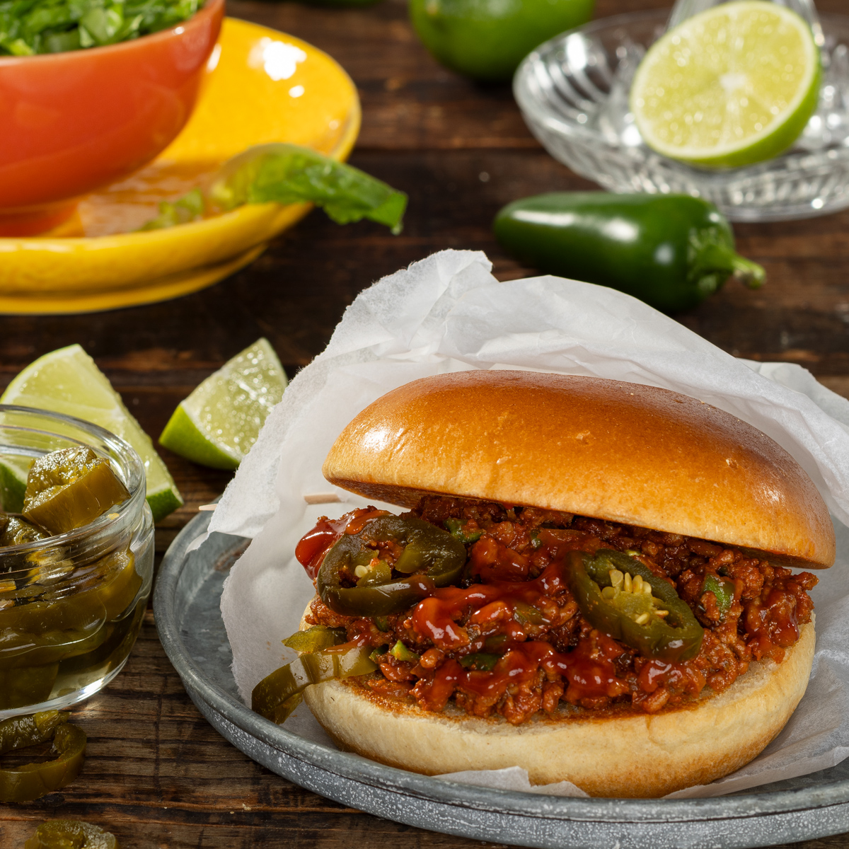 Im Vordergrund steht ein Sloppy Joe in einer offenen Tüte auf einem Glasschälchen. Im Hintergrund sind Limettenspalten und eine Jalapeno zu sehen.
