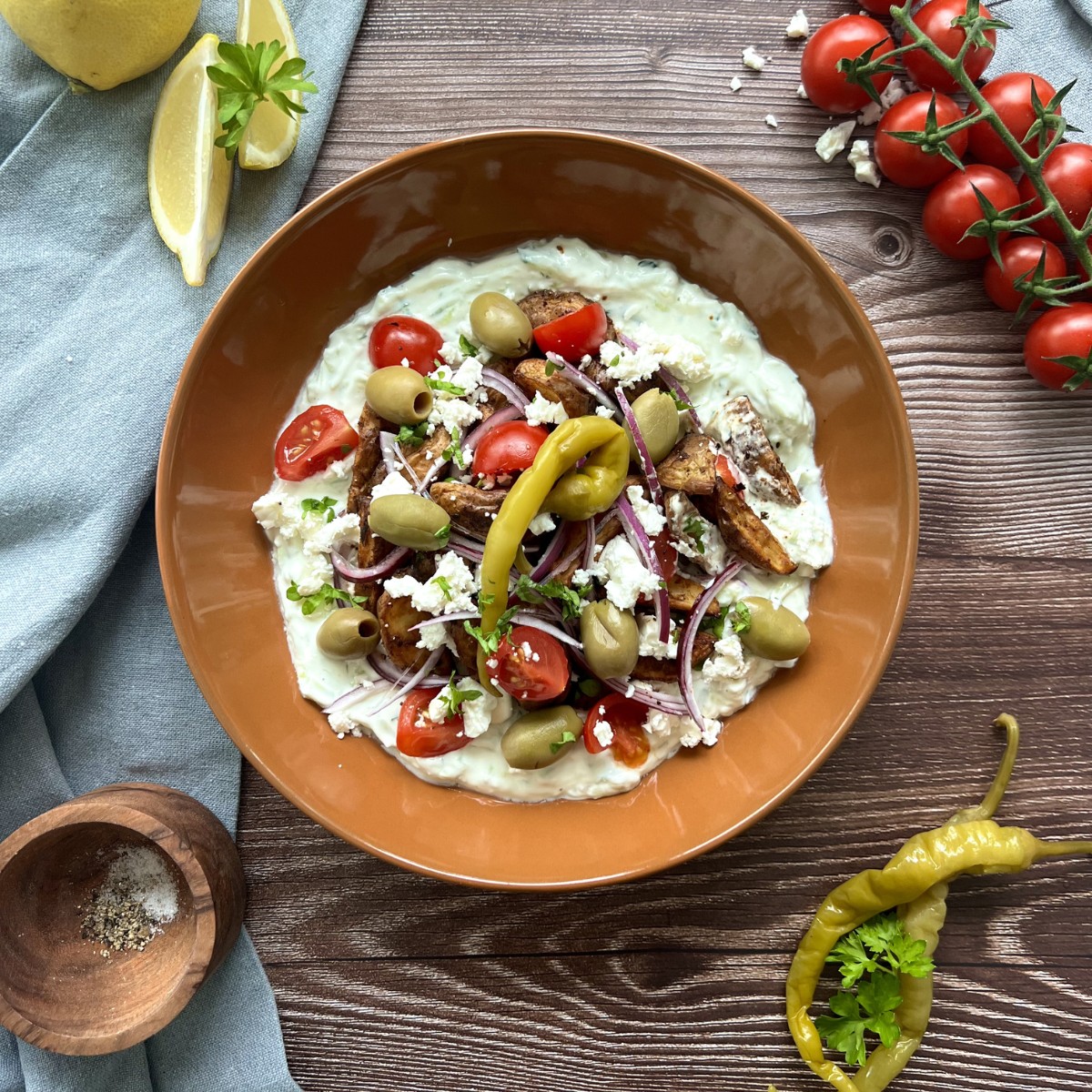 In der Mitte des Bildes steht die Bowl getoppt durch Oliven, Peperoni und Cherry Tomaten, neben der Bowl liegen Zitronenscheiben