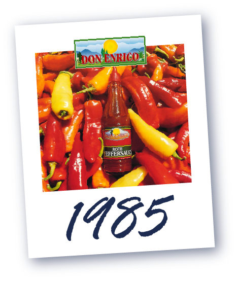 Flasche von Don Enrico mit roter Peffersauce, umgeben von roten und gelben Chilischoten, darunter die Jahreszahl '1985'.