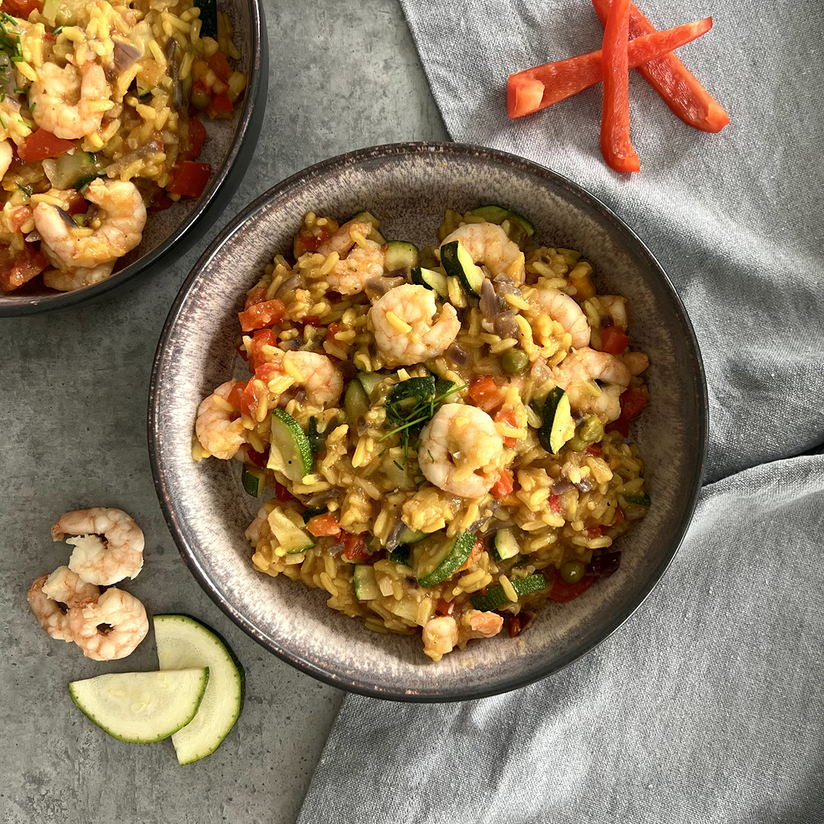 das Bild zeigt einen tiefen Teller geüllt mit Paella mit Garnelen, Zucchini und Paprika. Ein zweiter Teller wird angeschnitten.