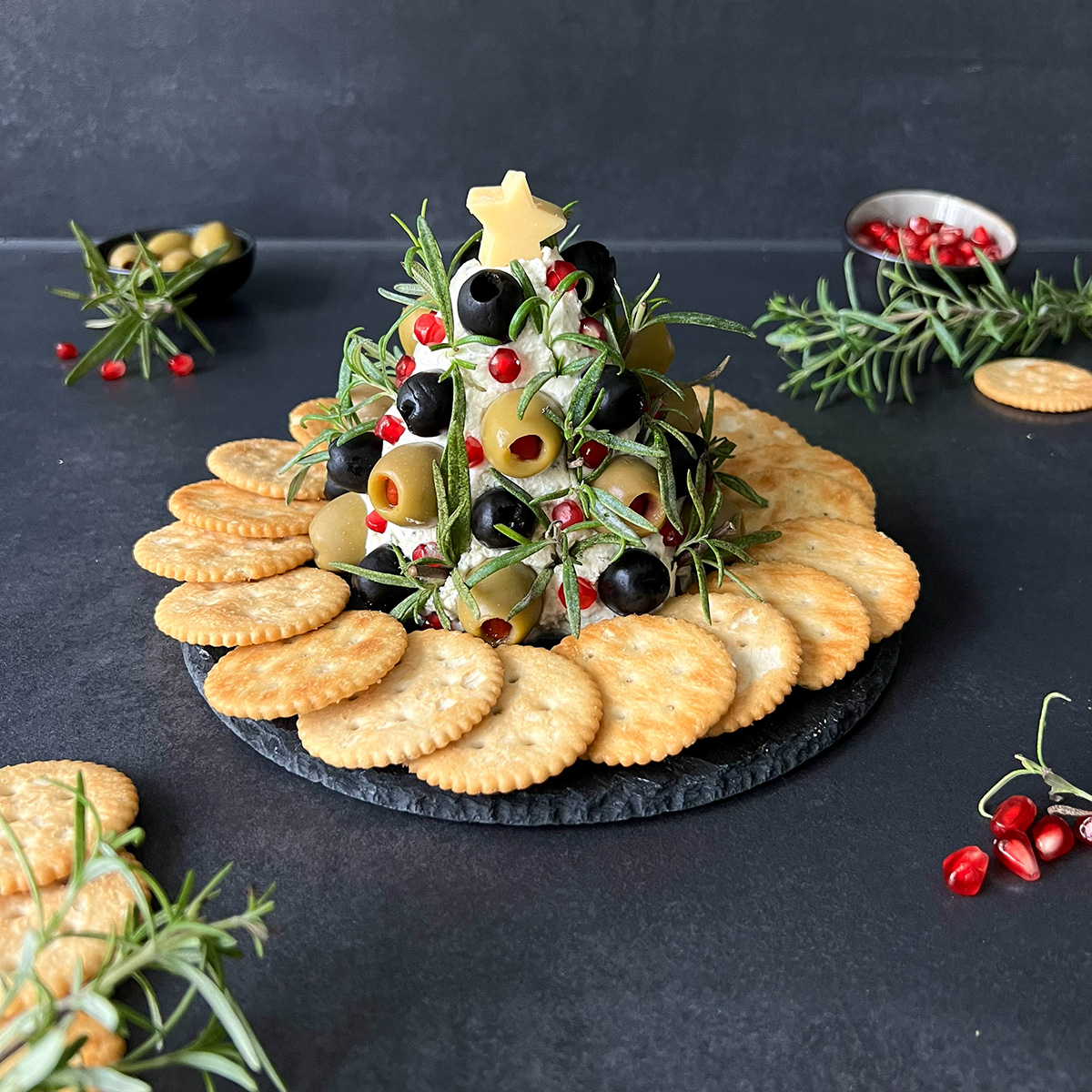 frischkäsetannenbaum mit Oliven auf einer Platte, daneben Rosmarin und Kekse