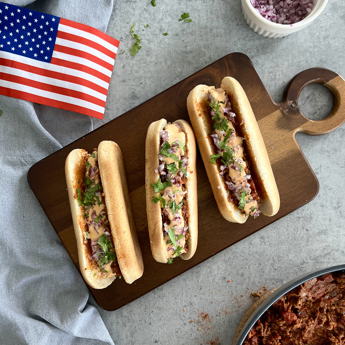 Das Bild zeigt drei Chili Cheese Hot Dogs auf einem Brett liegen. Daneben liegt eine US-amerikanische Flagge.
