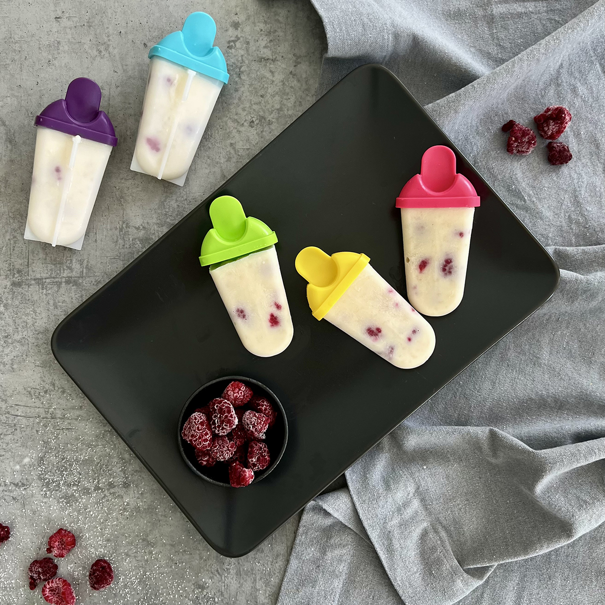 Das Bild zeigt 5 selbstgemachte Eis mit verschiedenen bunten Stäben. Es liegen Himbeeren zu Dekoration bereit.