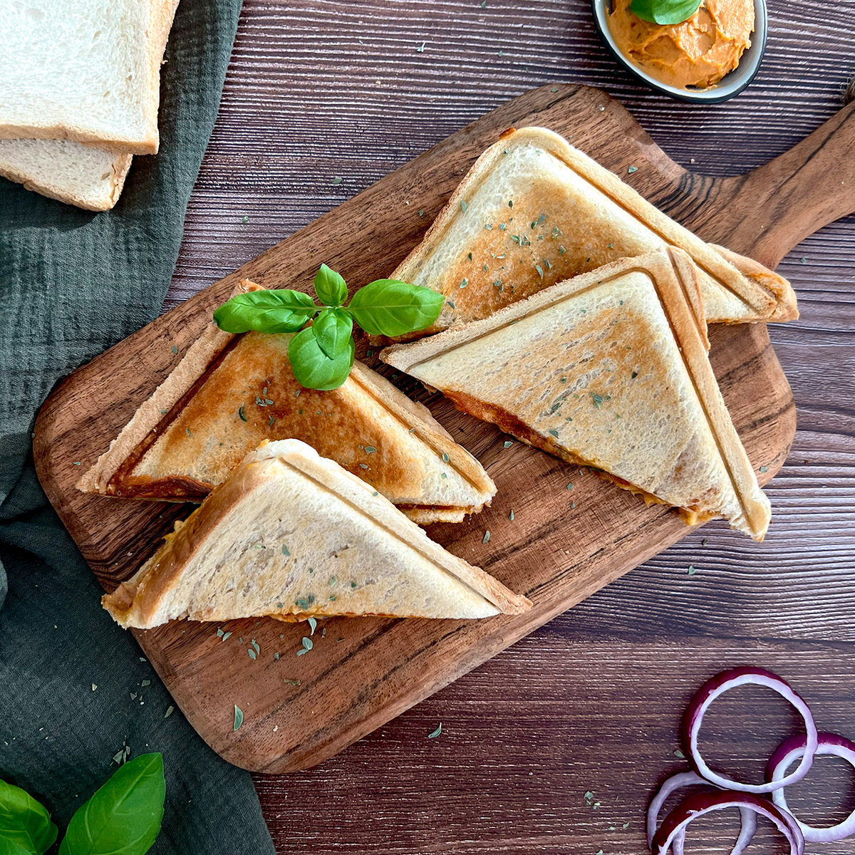 halbierte vegane Sandwiches auf einem Holzbrettchen getoppt mir Basilikum, daneben einzelne Zutaten wie Zwiebeln und Toast