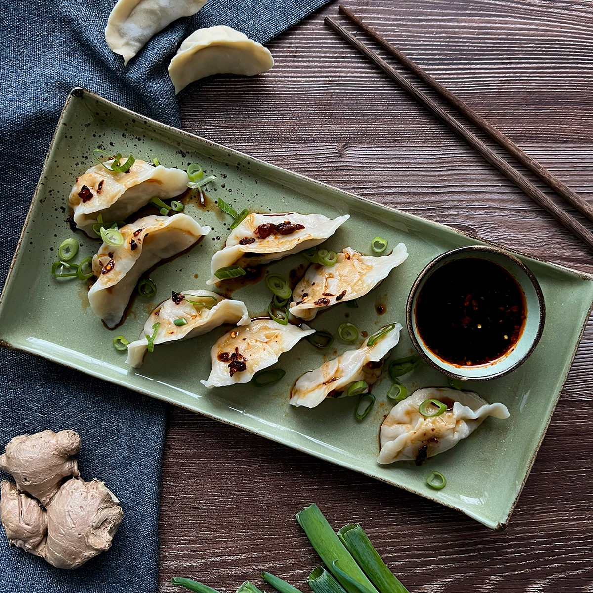 8 chinesisch gefüllte Teigtaschen auf einem grünen Teller mit Sojasauce als Dip, daneben einzelne Zutaten wie Ingwer und Frühlingszwiebeln