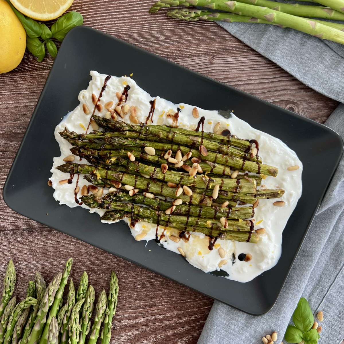 Auf einem länglichen schwarzen Teller ist Burrata verteilt und darüber liegt der grüne Spargel, getoppt mit Balsamico Creme und Pinienkernen.