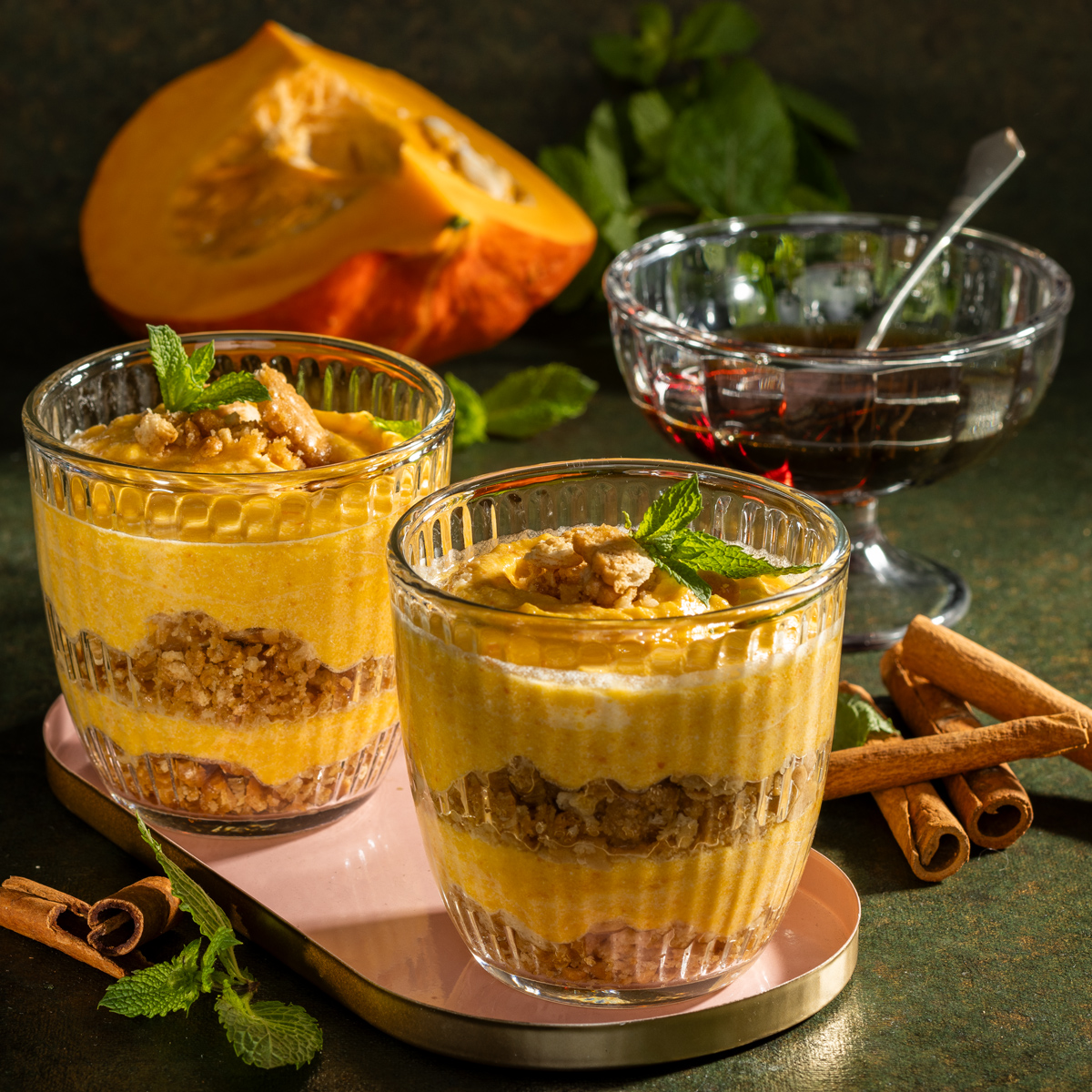 Zwei Gläser abwechselnd gefüllt mit einer Kürbiscreme und Haferflocken Masse. Im Hintergrund liegt ein Kürbis und Zimtstangen sind verteilt.