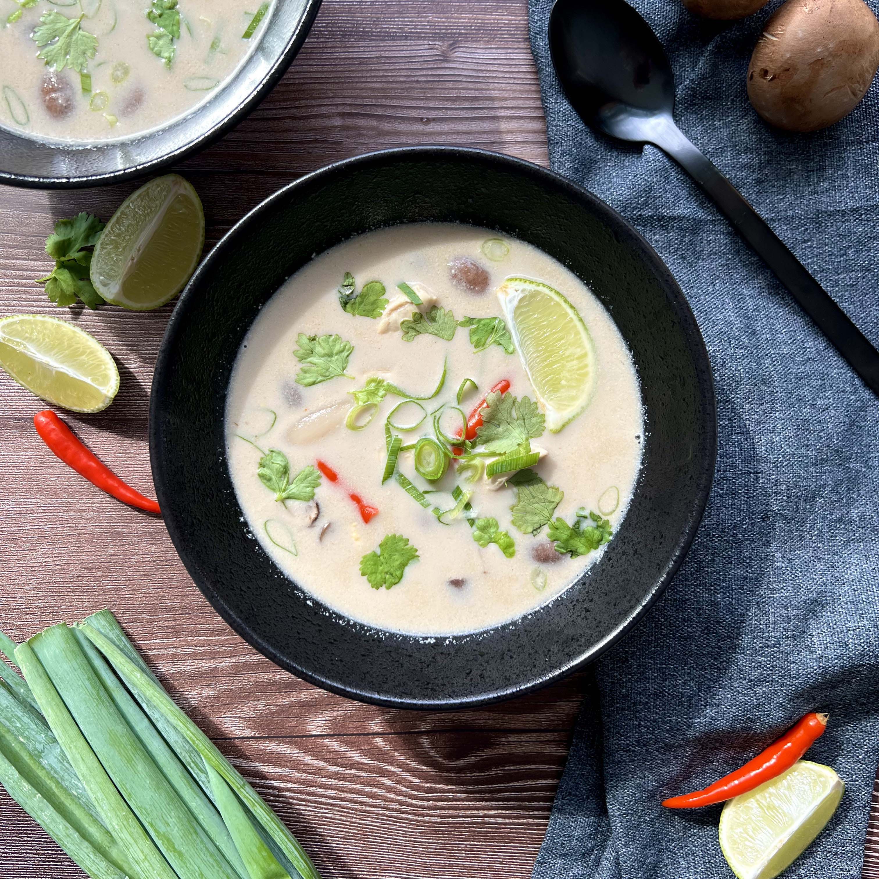 Das Bild zeigt einen schwarzen Teller gefüllt mit der Suppe. Limetten, Lauch, Pilze und Hähnchen sind darin zu sehen. Chili, Lauch und ein zweiter Teller sind angeschnitten.