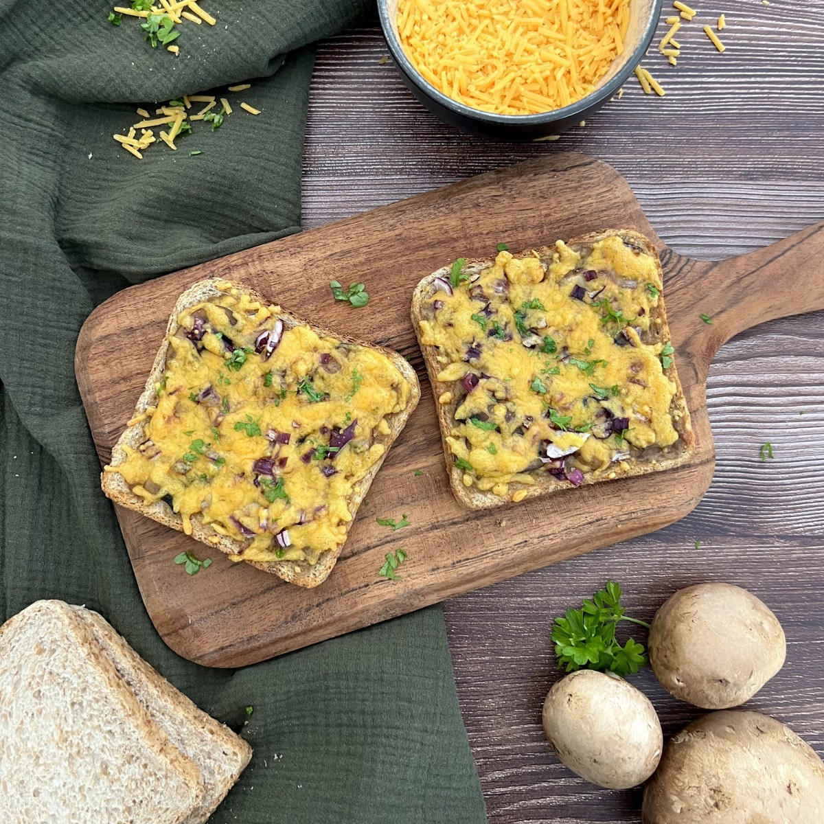 Zwei Champignons Toasts liegen auf einem Holzbrett. Die Toasts sind mit viel Käse und Petersilie getoppt.