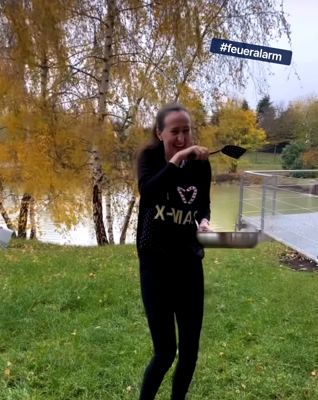 Eine Frau im schwarzen Pullover steht lachend auf dem Rasen und hält eine Pfanne in der Hand. 