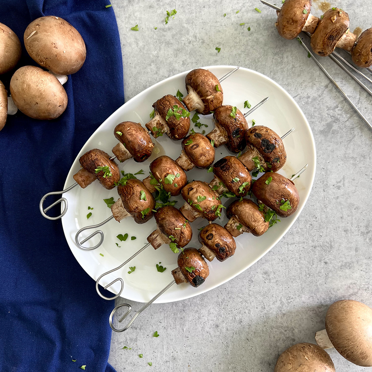 Knoblauch Balsamico Champignons Spieße Vier Champignonsspieße auf einem weißen Teller. Die marinierten Champignons sind im Ganzen und haben Grillspuren.