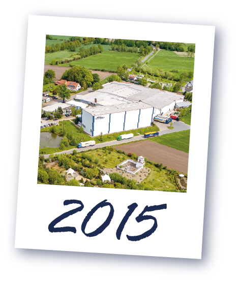 Luftaufnahme der großen weißen Logistikhalle umgeben von grünen Feldern und Bäumen, darunter die Jahreszahl '2015'.