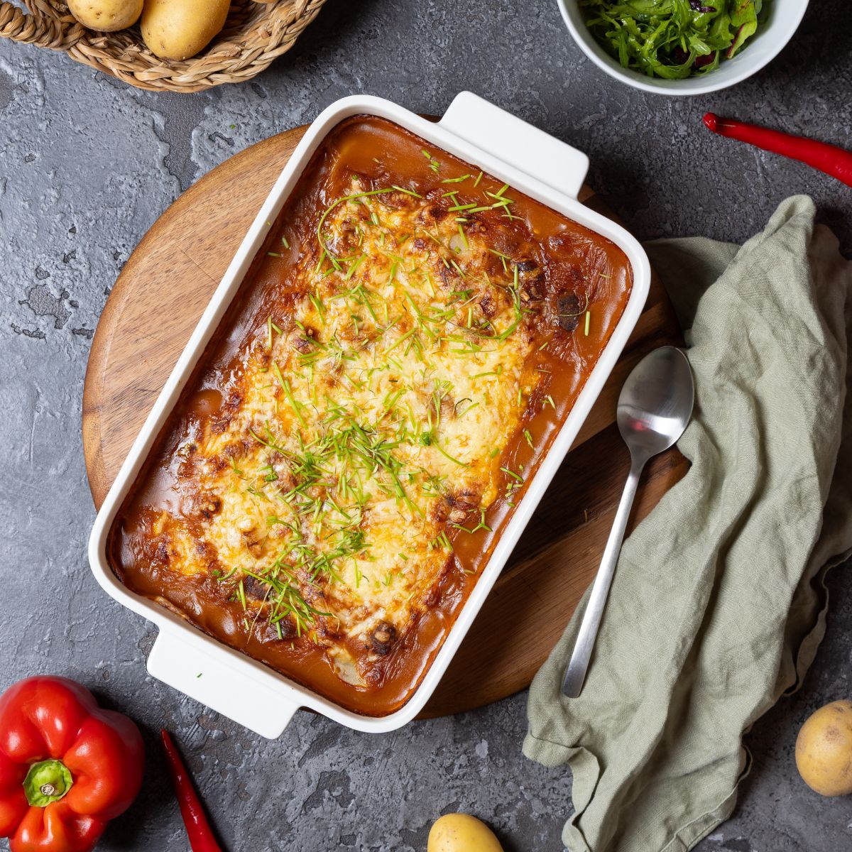Ein Auflauf in einer From getoppt mit Kräutern, daneben einzelne Zutaten wie Paprika oder Kartoffeln